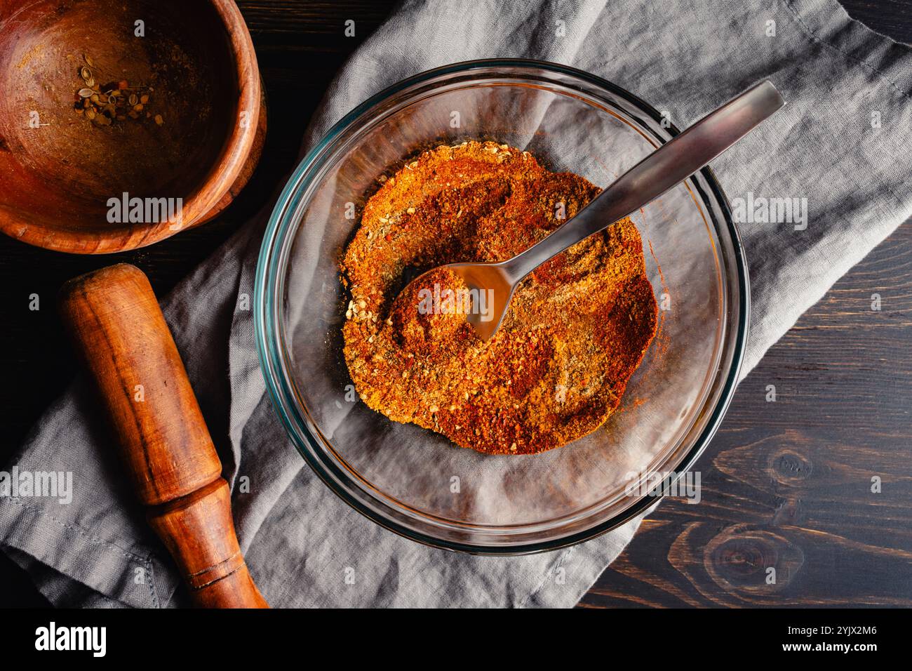 Mixing Spices to make Berbere Spice Blend: Ground herbs and spices mixed with a spoon in a glass bowl next to a wooden mortar and pestle Stock Photo