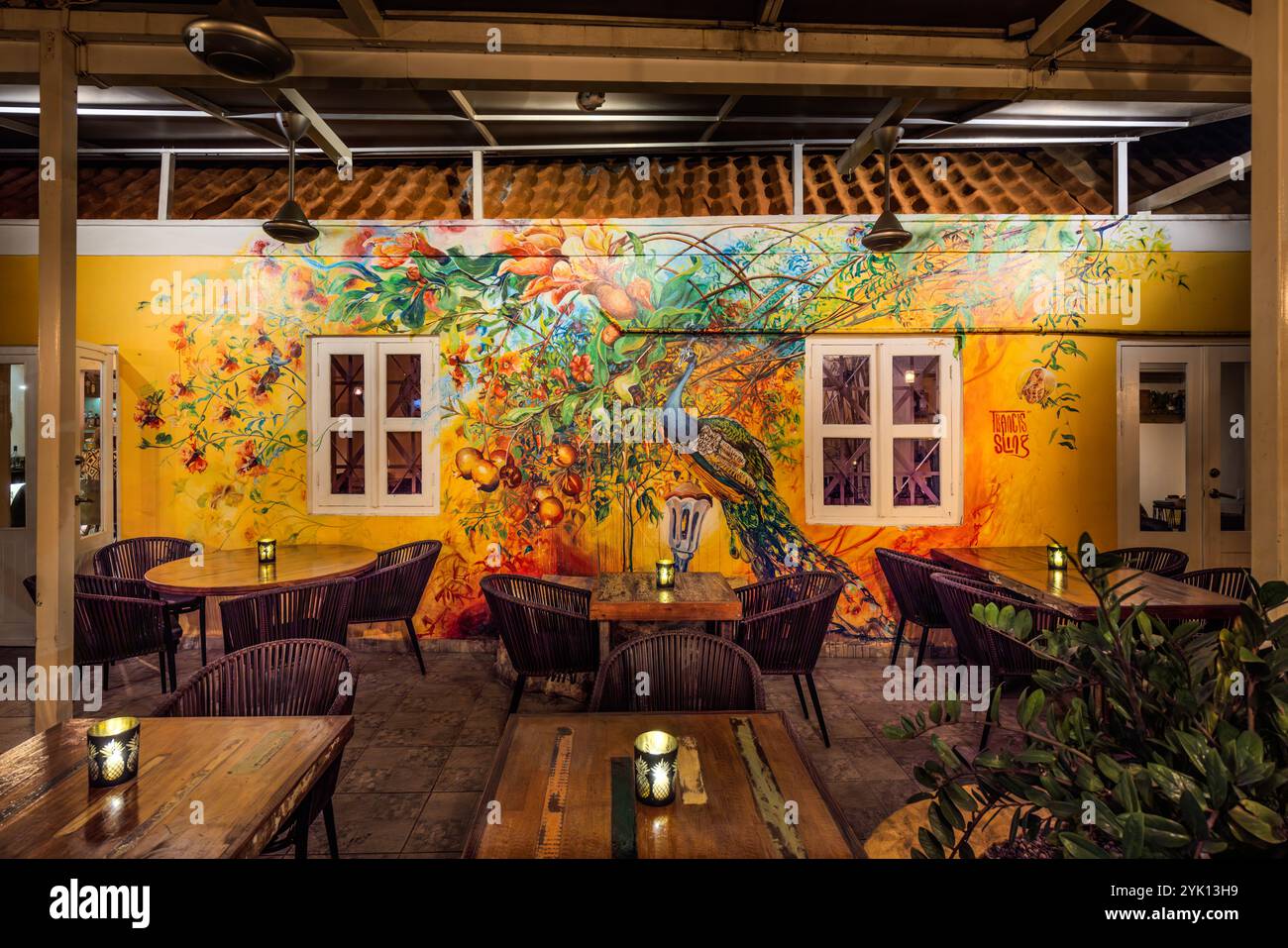 Inside MosaCaña Bar & Kitchen, Penstraat 41, Willemstad, Curaçao, Francis Sling's mural of a peacock and tropical fruits dominates the yellow back wall of the semi-open dining area. The interior of the old colonial building, with its terracotta-tiled roof, combines rustic wooden furniture with a street-art aesthetic; the bar serves exclusively rum, tequila and mezcal. Stock Photo