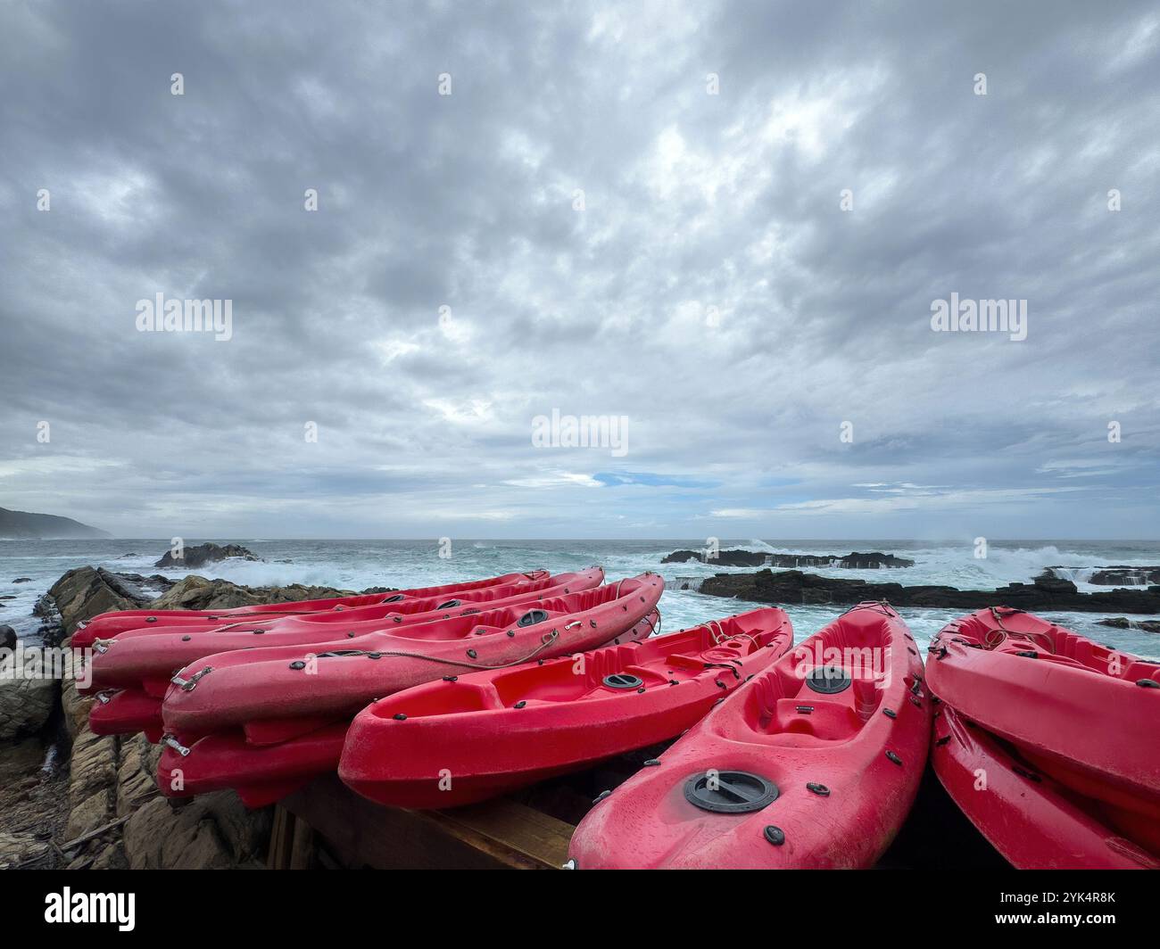Red kayaks stacked at the coast. Ocean waves crashing against the rocks, overcast sky, cloudy day by the sea Stock Photo