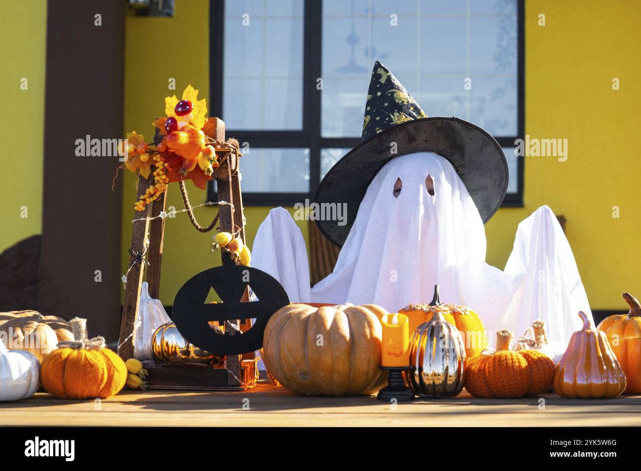 Child in bed sheet with slits like ghost in witch's hat and Halloween decor on the porch of the house outside in the yard of pumpkin, lantern, garland Stock Photo