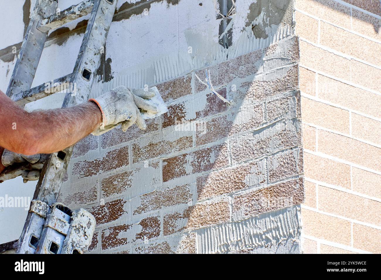 Installing thin decorative and protective panels with imitation brickwork on facade of house, house painter uses steel putty knife to smooth out glued Stock Photo