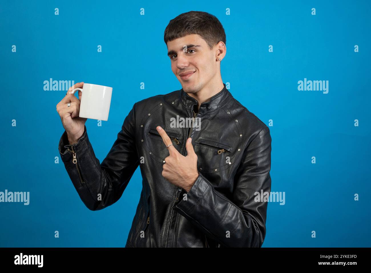 Young man wearing black leather jacket holding and pointing at white mug on blue background Stock Photo