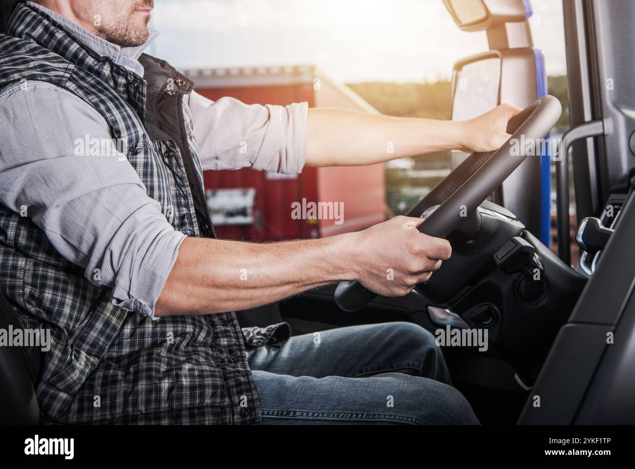 A truck driver is focused on steering his vehicle, parked alongside large shipping containers during the golden hour of sunset. Stock Photo