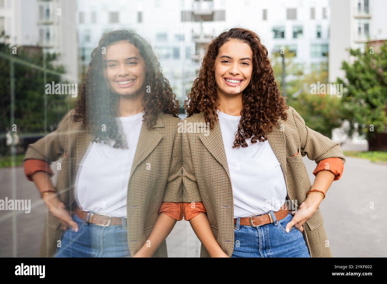 A cheerful Brazilian woman in business attire poses confidently with her reflection in a glass pane in an urban landscape, epitomizing modern, empower Stock Photo
