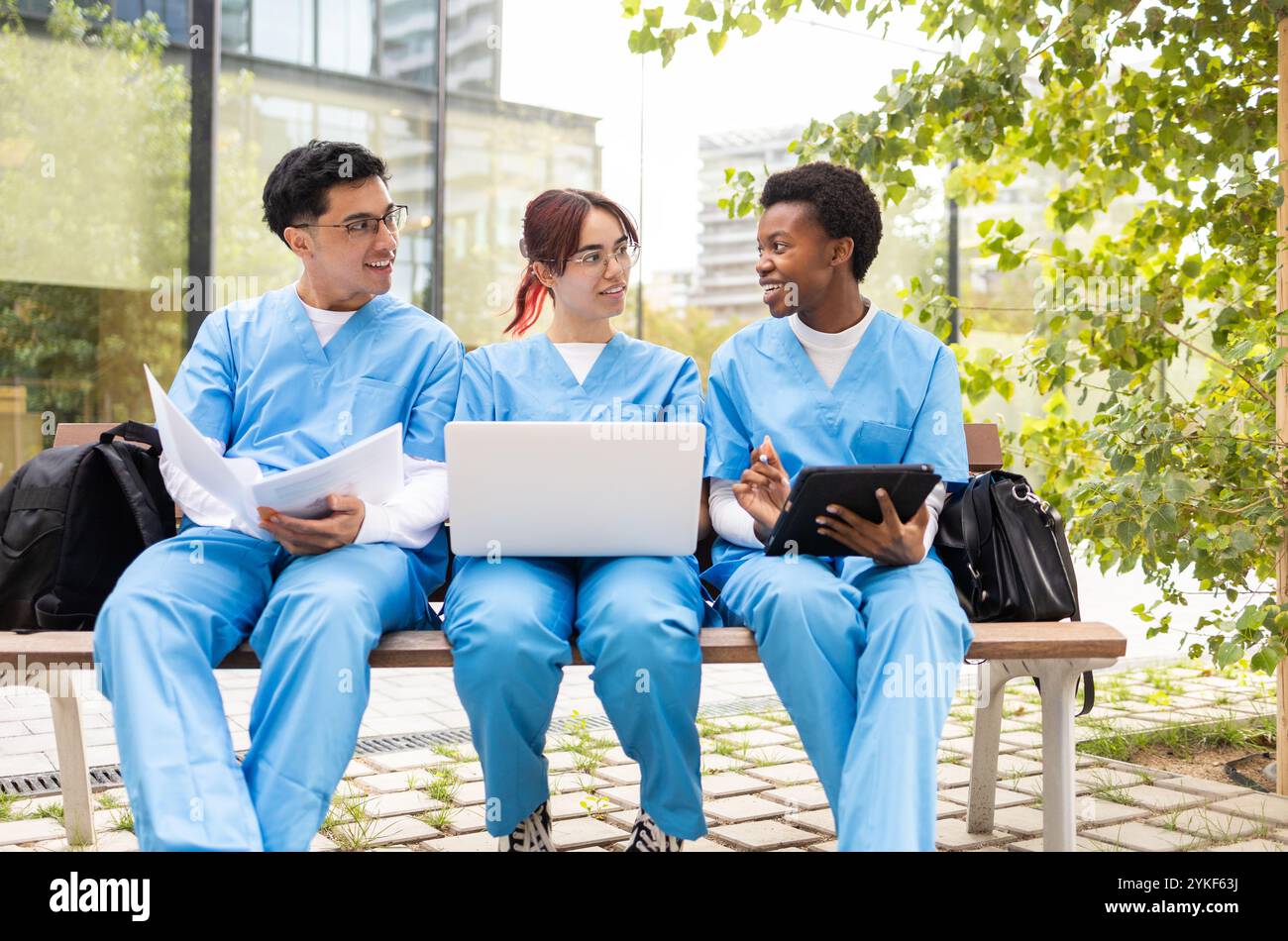 Three nursing students in blue scrubs are engaged in a study session outside, using a laptop and digital tablets, surrounded by lush greenery Stock Photo