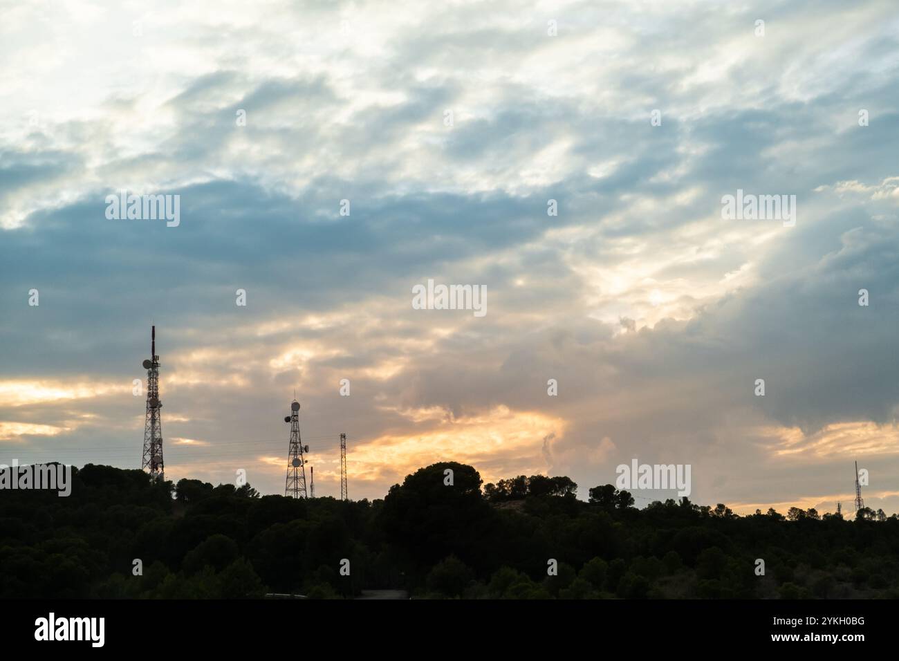 Cloudscape at sunset with many communication towers in silhouette over the sky. The horizon line is made of trees and the sky is threatening and storm Stock Photo