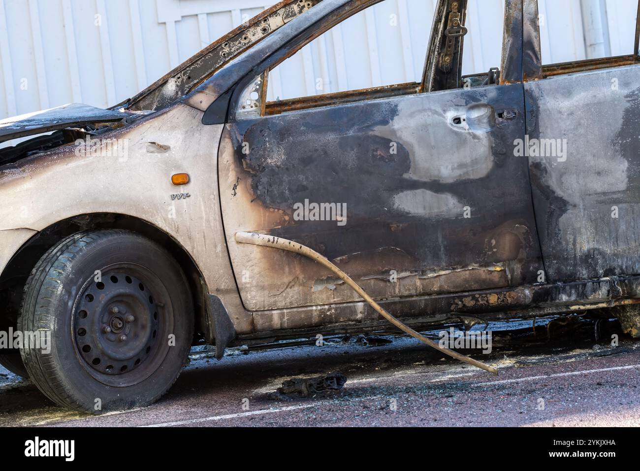 A burned-out car sits on the roadside, revealing severe fire damage. The charred exterior and melted parts reflect neglect and the aftermath of a fire Stock Photo