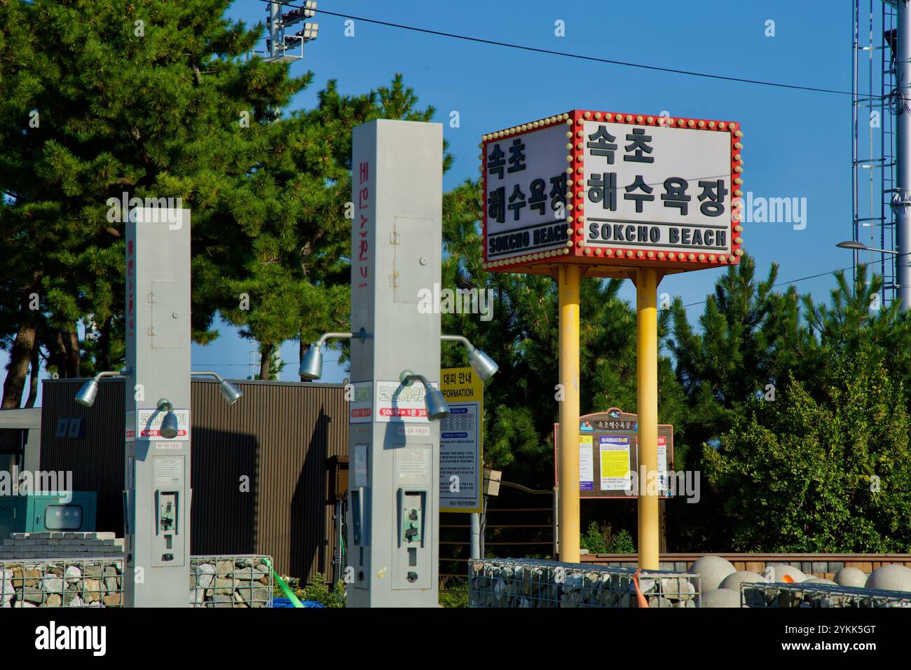 Sokcho, South Korea - November 3rd, 2024: Coin-operated outdoor shower columns stand near the prominent Sokcho Beach sign, surrounded by pine trees an Stock Photo