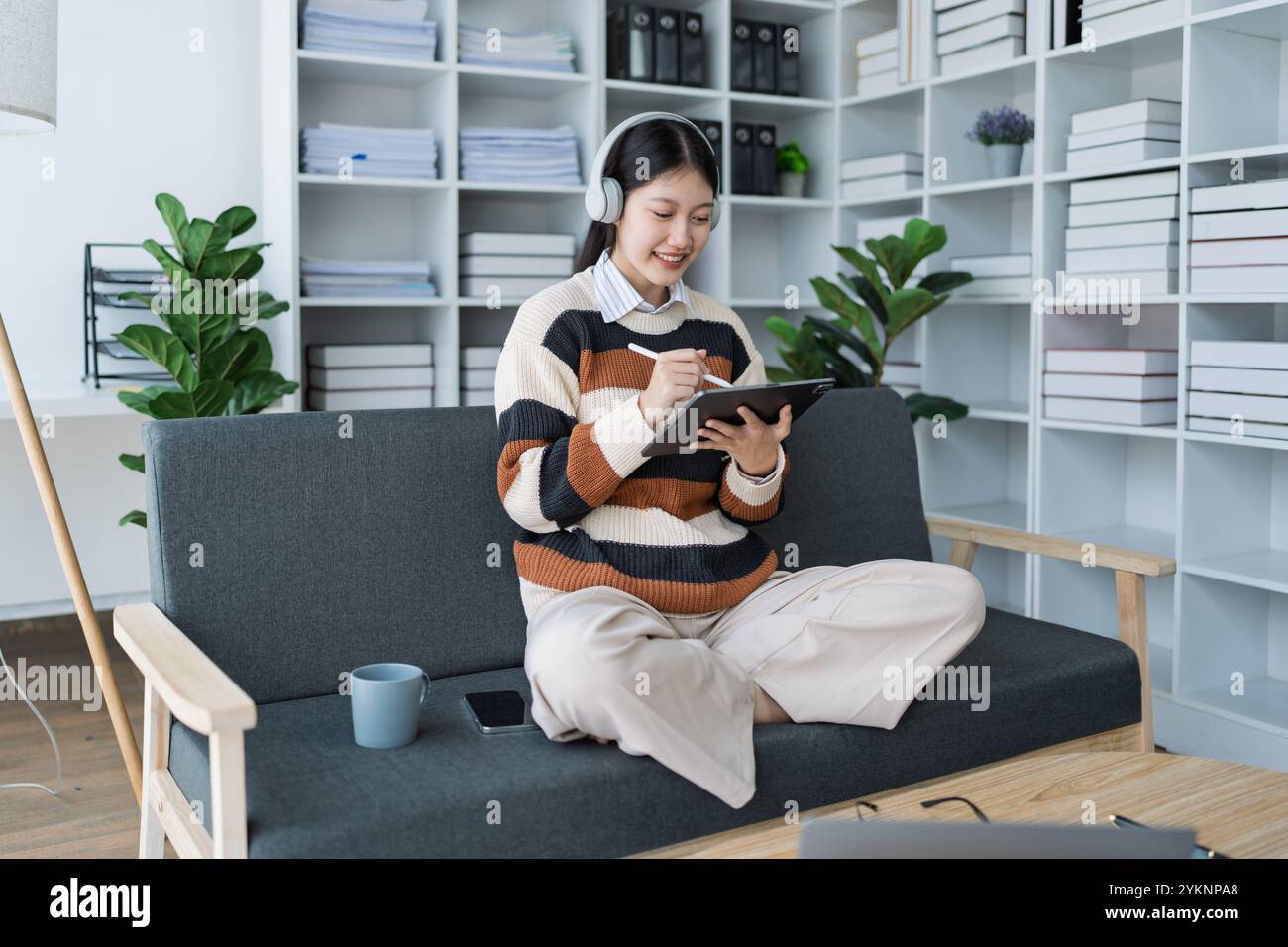 A young woman enjoys a moment of creativity as she uses her tablet on a cozy couch in her modern living room, surrounded by greenery and organized she Stock Photo