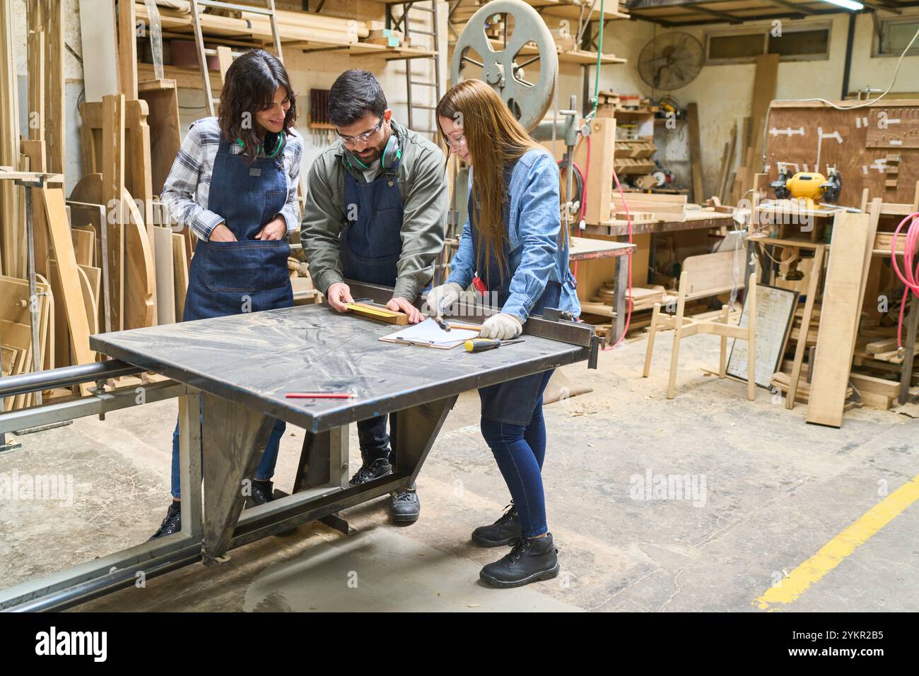 A group of young apprentices working together in a lumberyard workshop. Guided by an older mentor, they measure materials, emphasizing teamwork and le Stock Photo