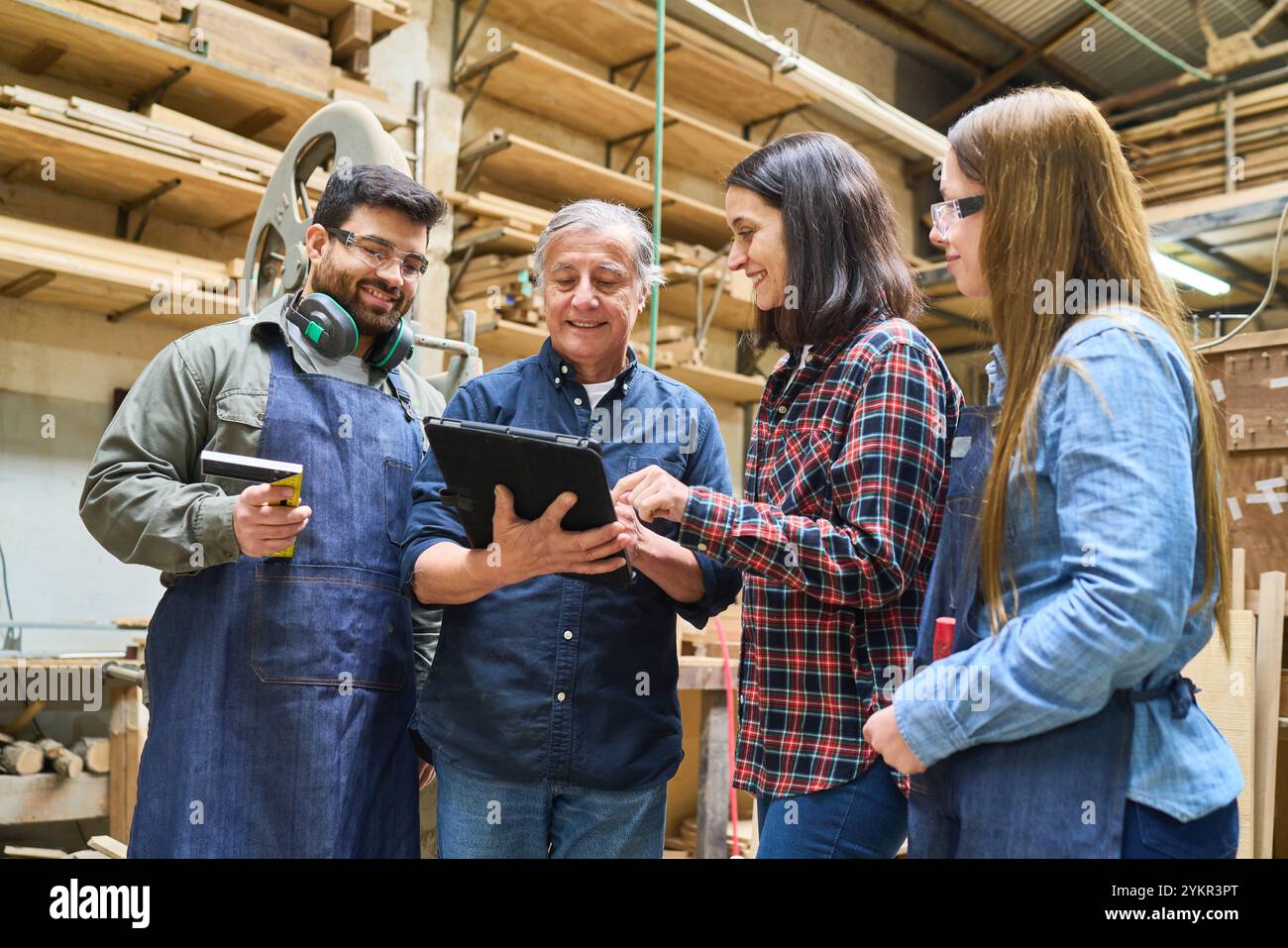 A group of young workers engage with an experienced mentor in a lumberyard. They are collaborating and learning hands-on woodworking skills, emphasizi Stock Photo