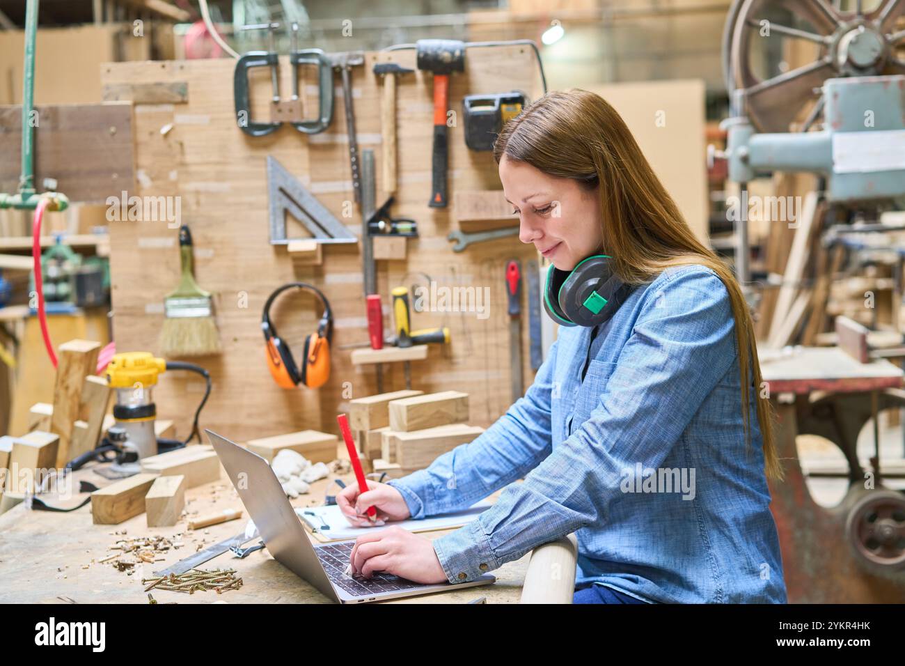 A young woman uses a laptop in a lumberyard, surrounded by tools and wood. The scene emphasizes focus, craftsmanship, and industrial work environments Stock Photo