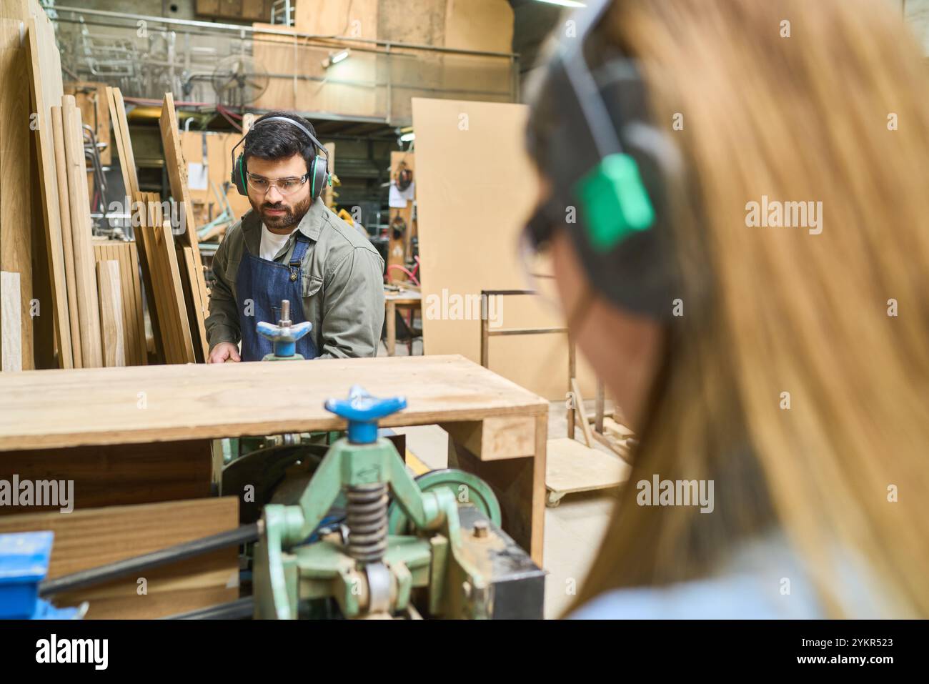 A young apprentice watches intently as a skilled worker operates machinery in a busy lumberyard, demonstrating teamwork and training in a hands-on env Stock Photo