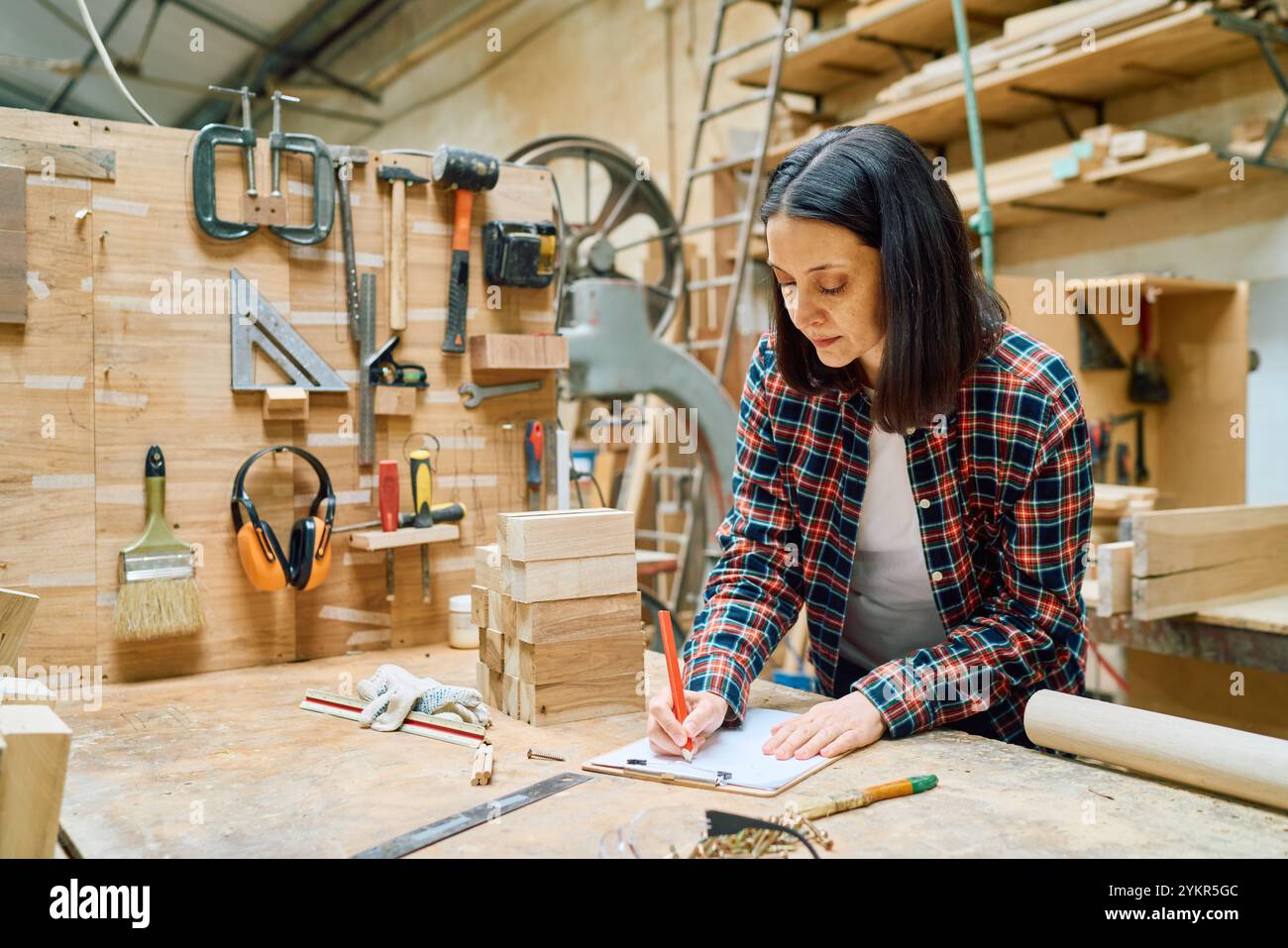 A woman concentrates on her task in a bustling woodworking shop. Surrounded by tools and wood, she epitomizes craftsmanship and dedication in a creati Stock Photo