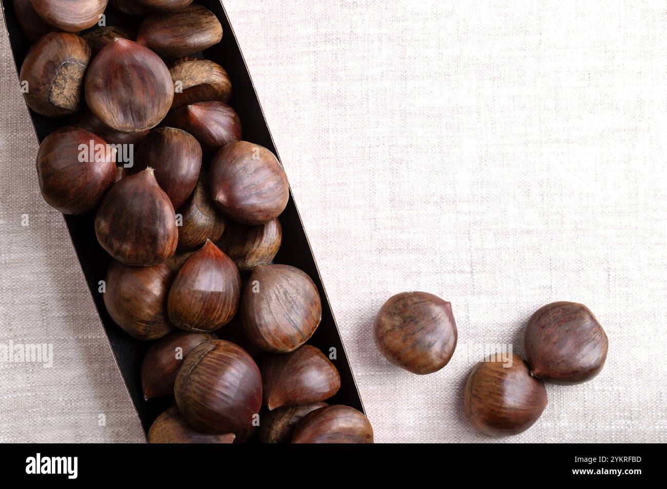 Sweet chestnuts in cardboard punnet on linen, from above, with empty space for text. Castanea sativa, raw, whole and unshelled. Stock Photo