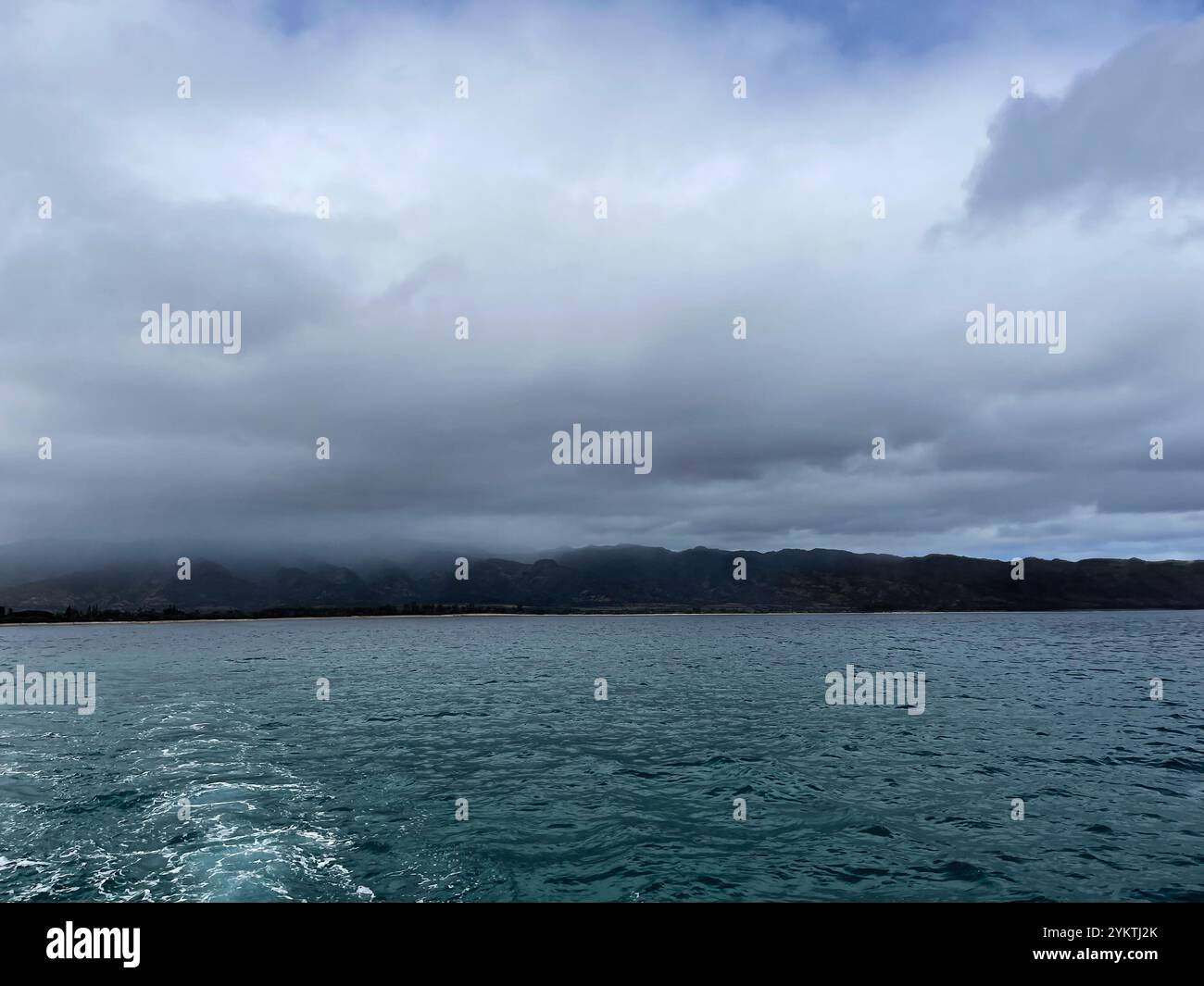 Looking back as you sail away from a tropical hawaiian island. The clouds and fog are rolling over the mountains. It's cloudy and misty. The water is - Smartphone Captured Stock Image