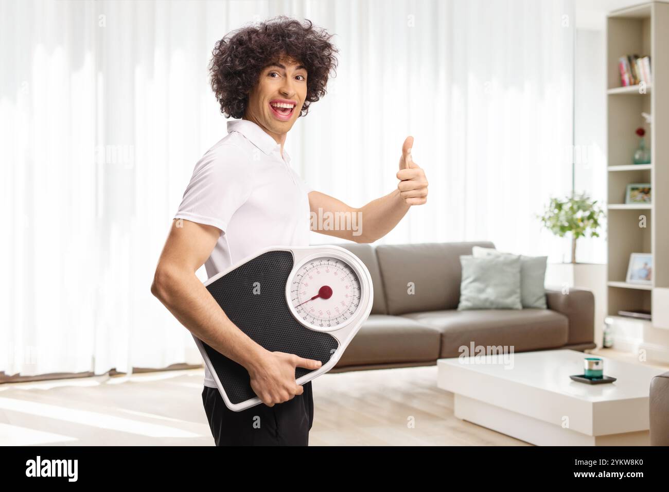 Young man holding a weight scale at home and gesturing a thumb up sign ...