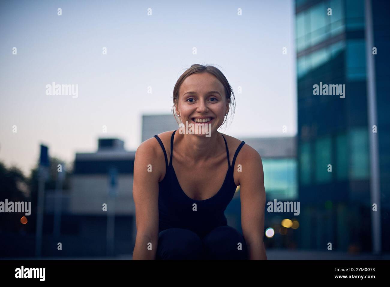 Young woman smiling while practicing yoga outdoors at sunset near modern buildings Stock Photo
