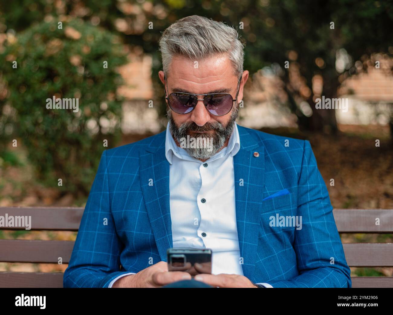 A hipster mature business man using mobile phone app texting while sitting on the park bench.  Stock Photo
