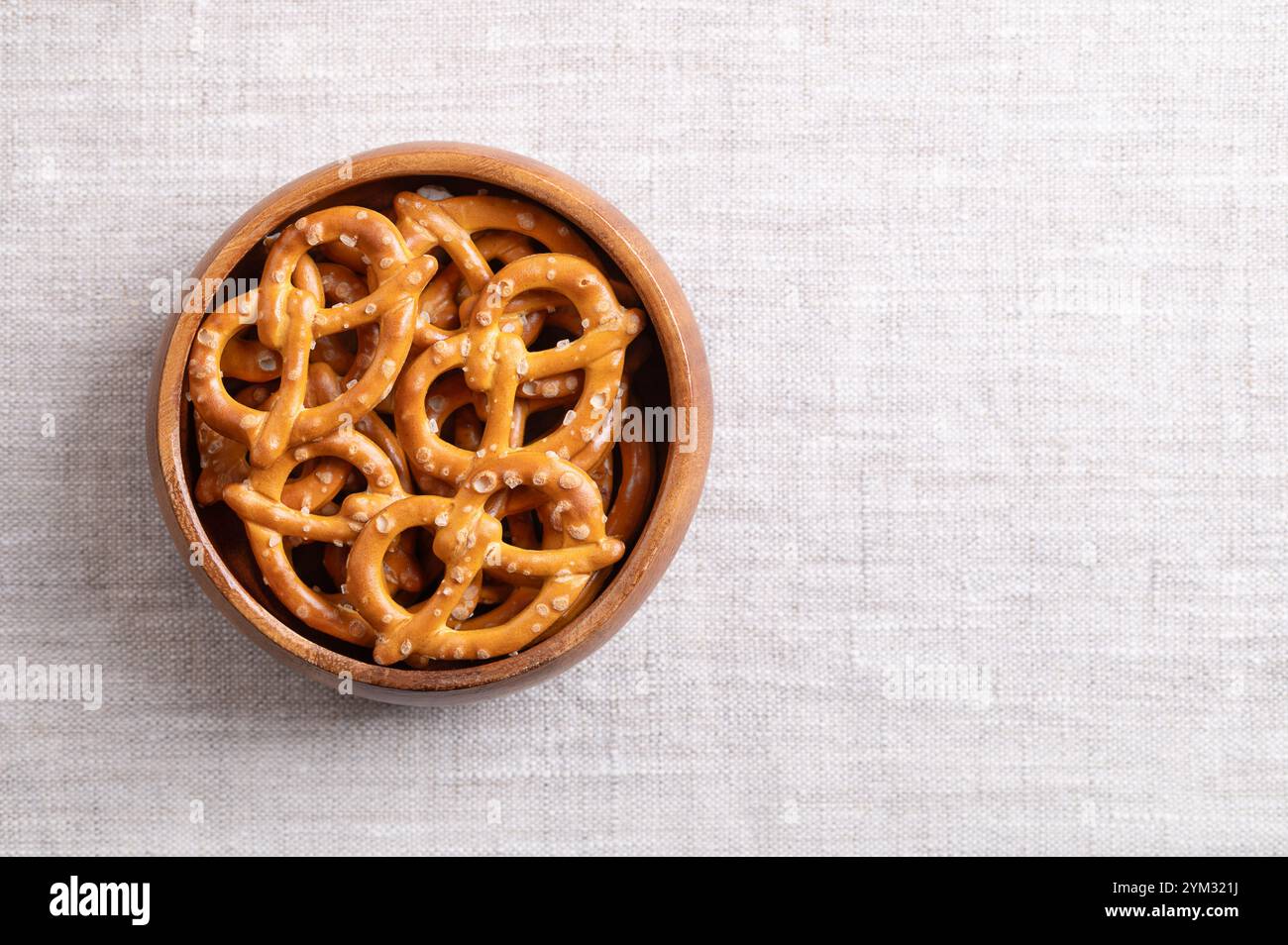 Salted pretzels in a wooden bowl on linen fabric, with empty space for text. Golden toasted small pretzels, in the traditional shape of a loop. Stock Photo