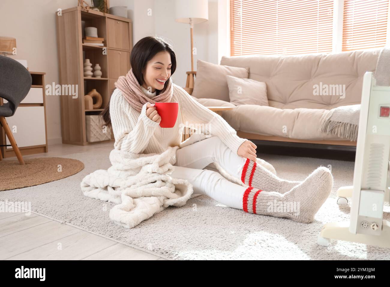 Young woman with cup of tea warming near radiator at home on winter day ...