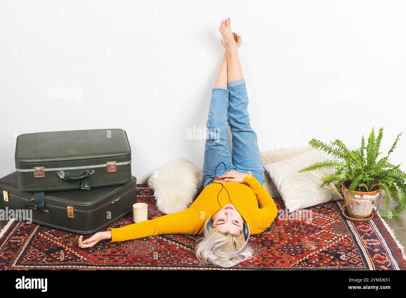 Caucasian woman with blonde hair wearing a yellow sweater and jeans, lying on a patterned rug, listening to music with headphones, legs up on the wall Stock Photo