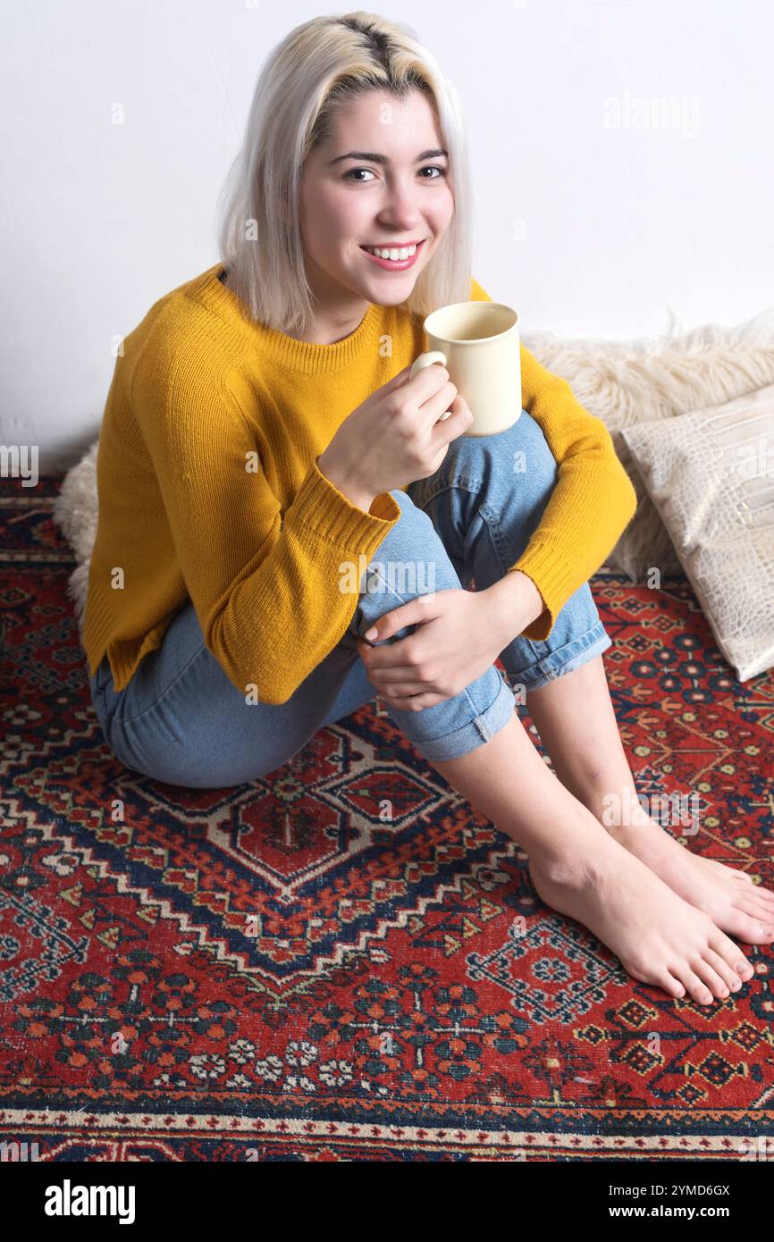 Caucasian woman with blonde hair, in a yellow sweater and jeans, smiling warmly while holding a mug. Relaxed and cozy atmosphere on a patterned rug. Stock Photo