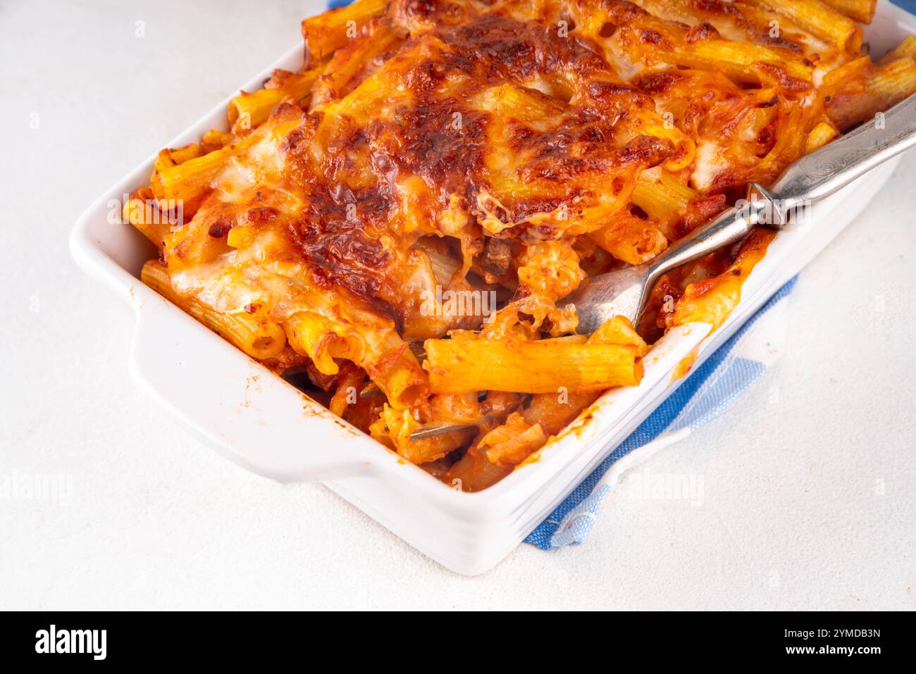 Baked Ziti pasta with bolognese or marinara tomato sauce and cheese, in baked pan and plate, on white kitchen table copy space Stock Photo