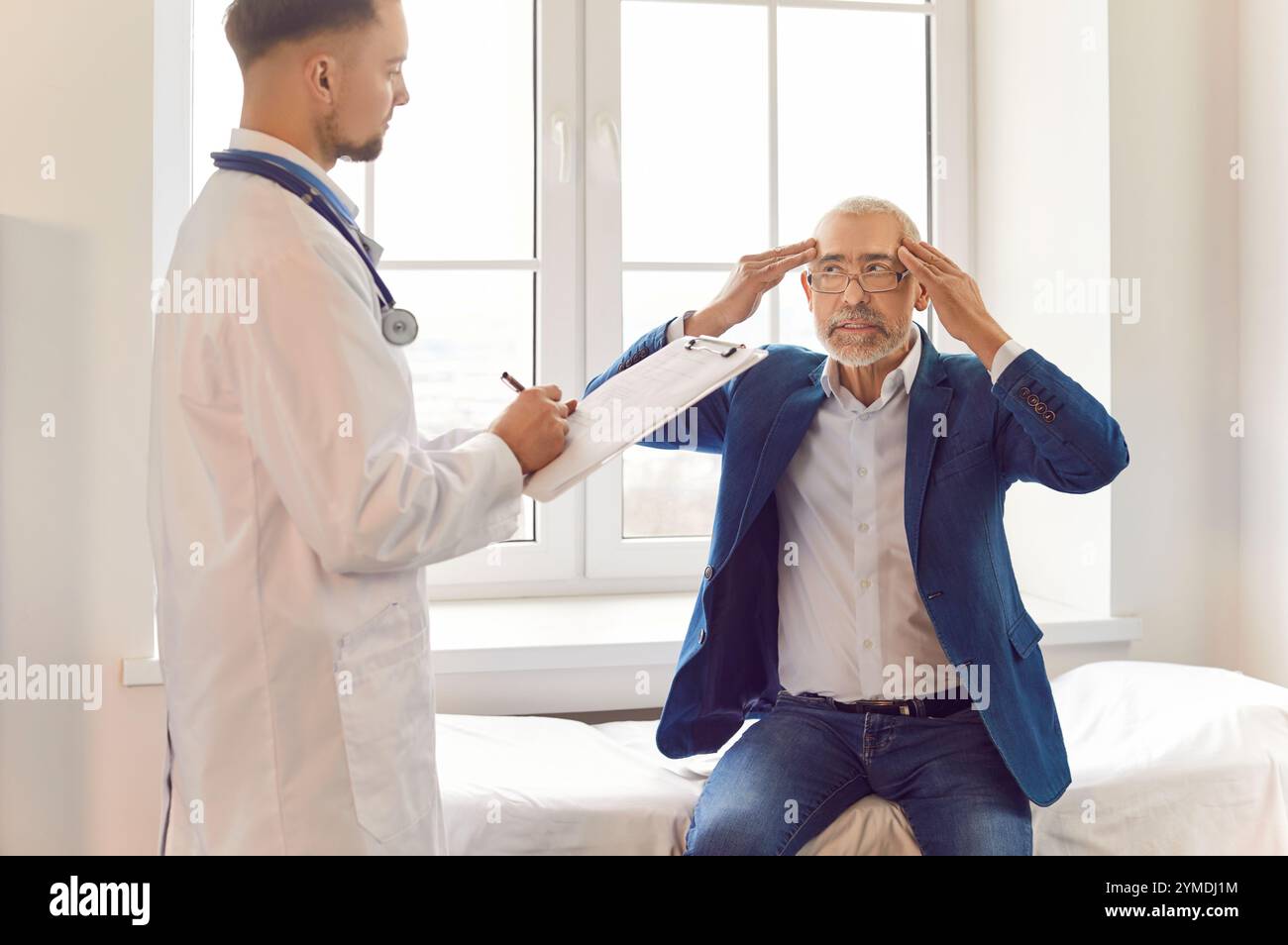 Sick senior man sitting on couch at doctor's office suffering from headache or having migraine. Stock Photo