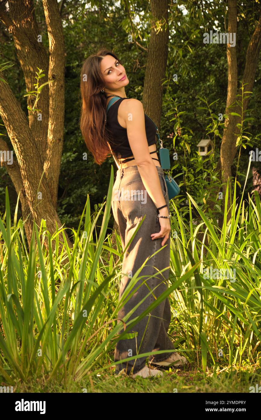Woman standing in a sunlit forest, wearing a black top and jeans ...