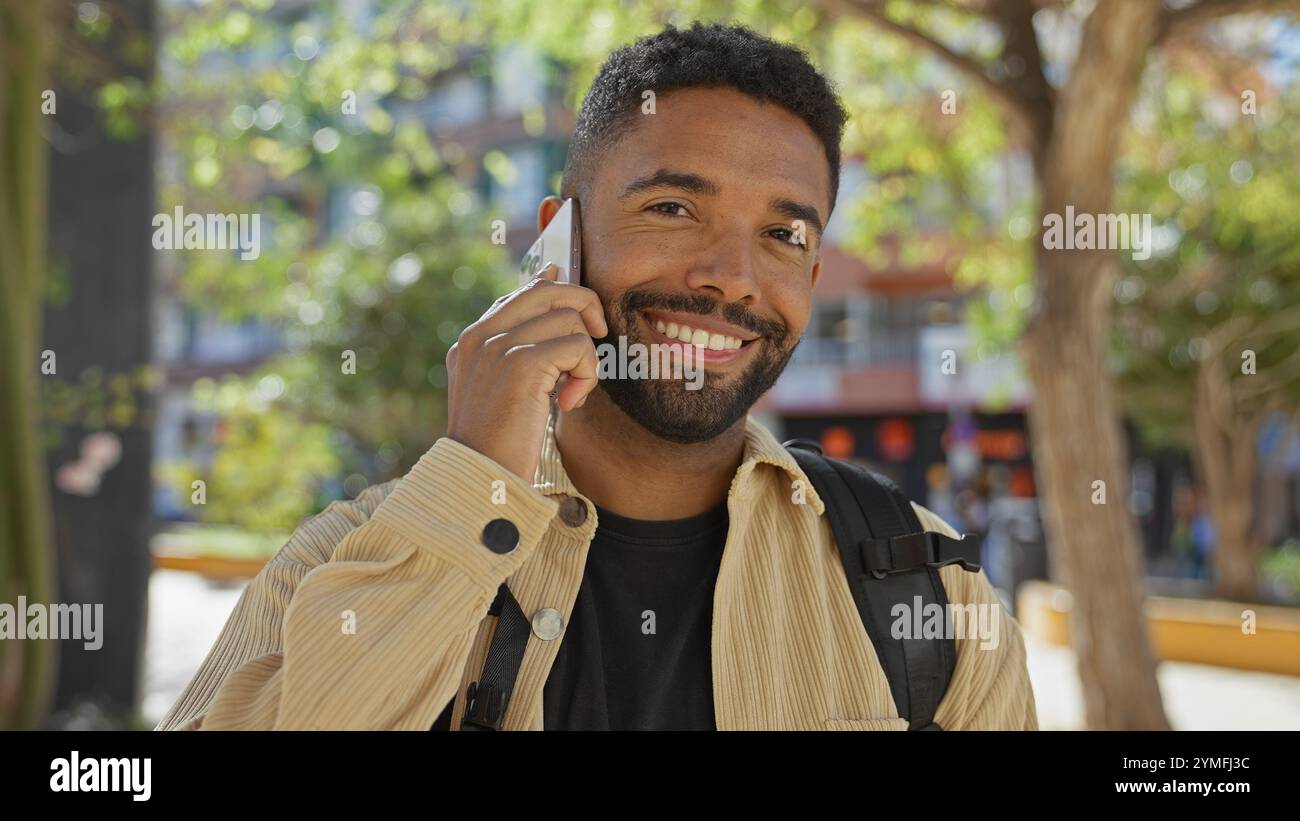A smiling young black man with a beard engaging in a phone conversation outdoors in an urban park setting. Stock Photo