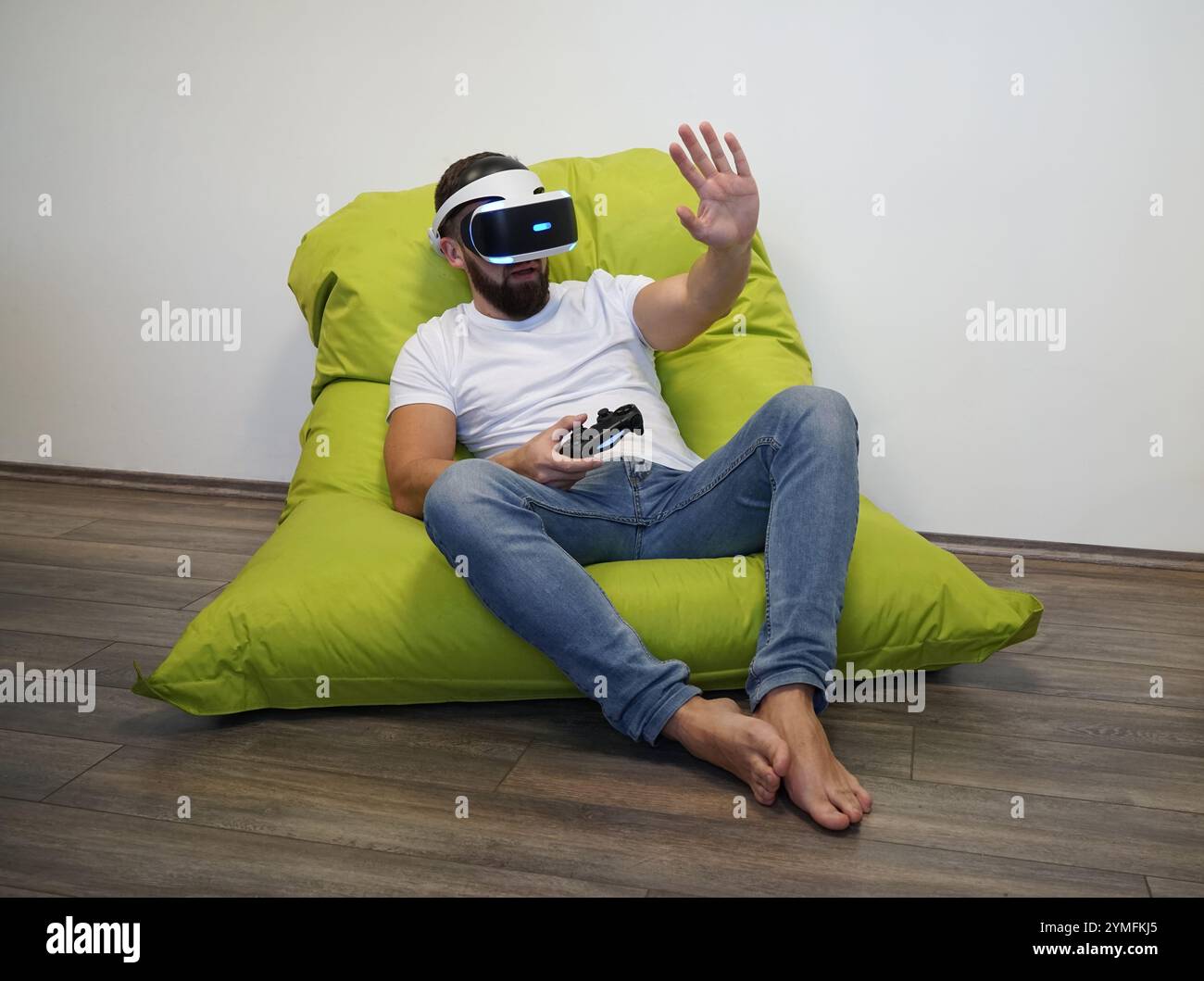 Man is seated on green bean bag, using VR headset and holding controller Stock Photo