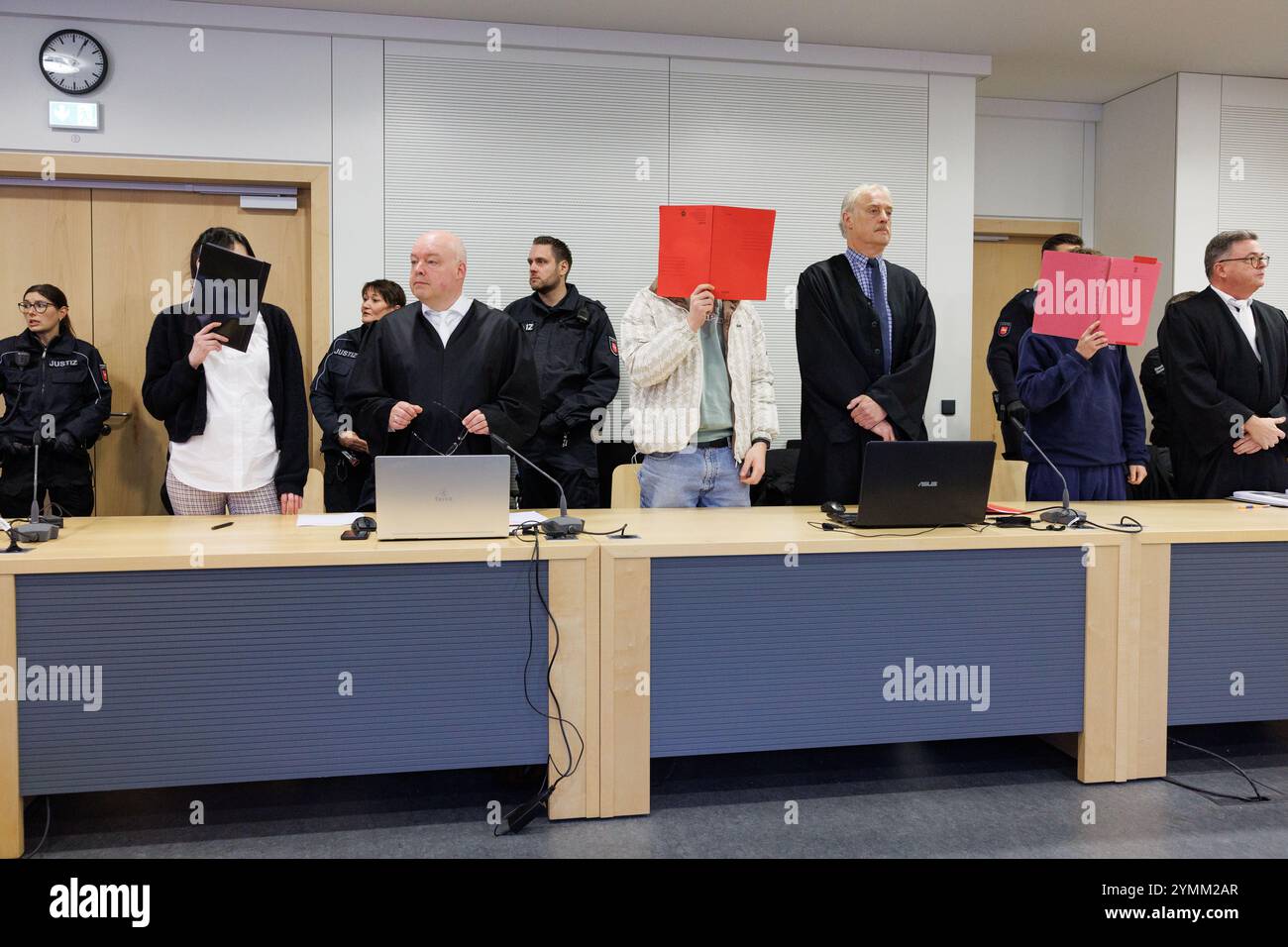 22 November 2024, Lower Saxony, Osnabrück: Three defendants, their faces covered with folders, stand next to their defense lawyers in a courtroom at Osnabrück District Court. The public prosecutor's office is accusing the men, now aged 23 and 25, and the woman, now 27, of attempted joint manslaughter, dangerous bodily harm, extortion and robbery. They are said to have lured a 17-year-old girl into a forest near Meppen, assaulted her there and tied her up. The teenager was abused and injured for hours. Photo: Friso Gentsch/dpa Stock Photo