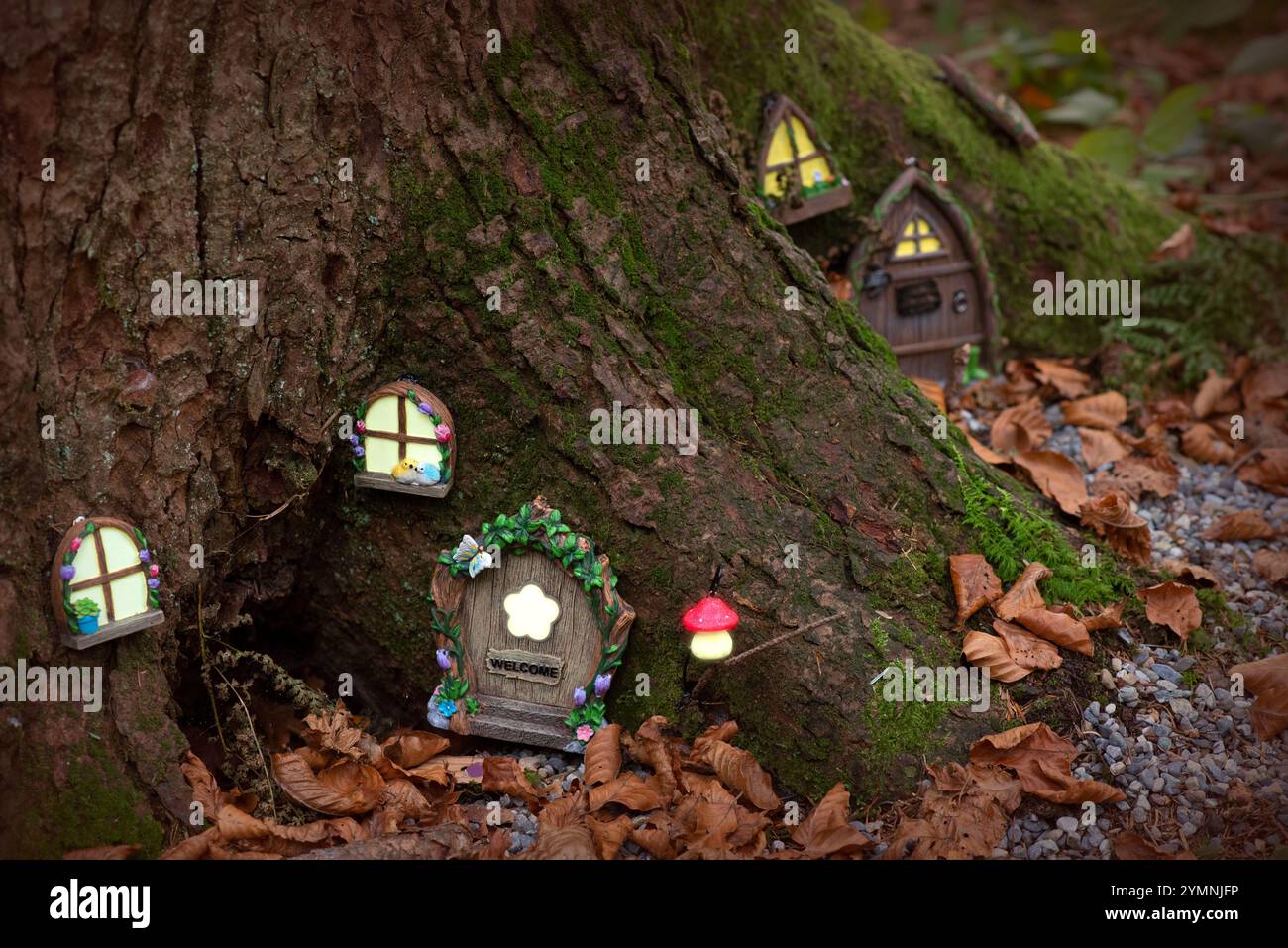 A whimsical forest scene featuring a tiny fairy-tale village carved into the roots of a large tree. The village includes glowing windows with floral d Stock Photo