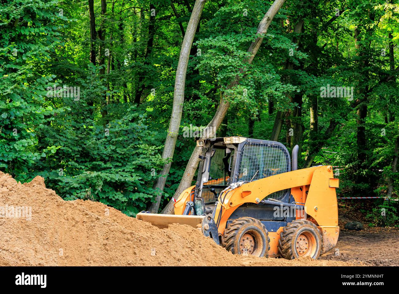 An orange skid steer loader shovels soil against a backdrop of trees. Stock Photo