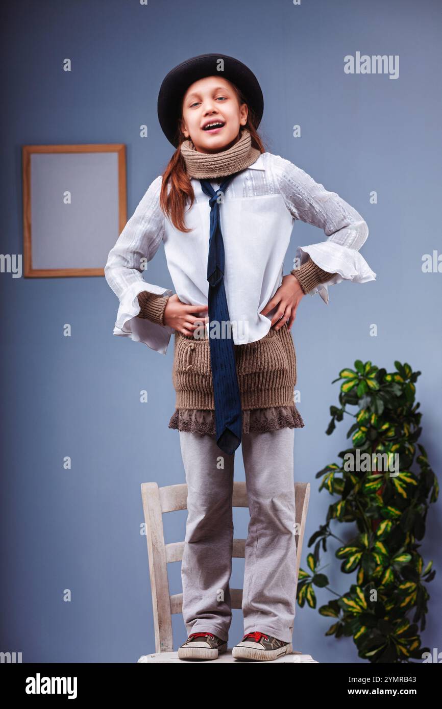 Young girl standing on a chair wearing an oversized man's tie and hat, hands on her hips, looking at the camera and smiling Stock Photo