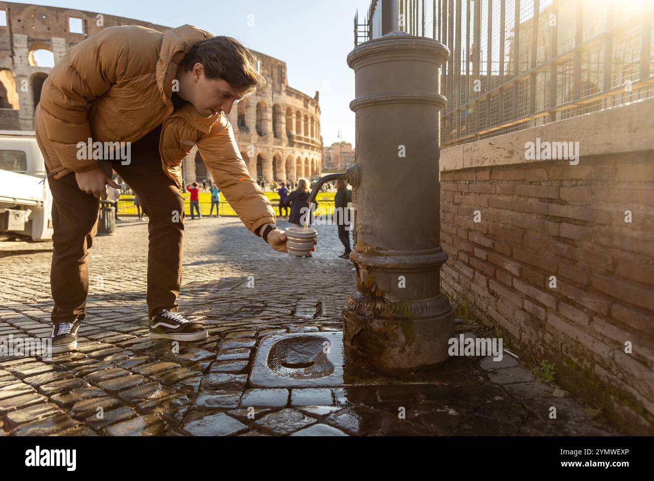 Rome, Italy - Nov 15, 2024: A tourist fills a cup at a nasone, a free public fountain, with the Colosseum in the background Stock Photo