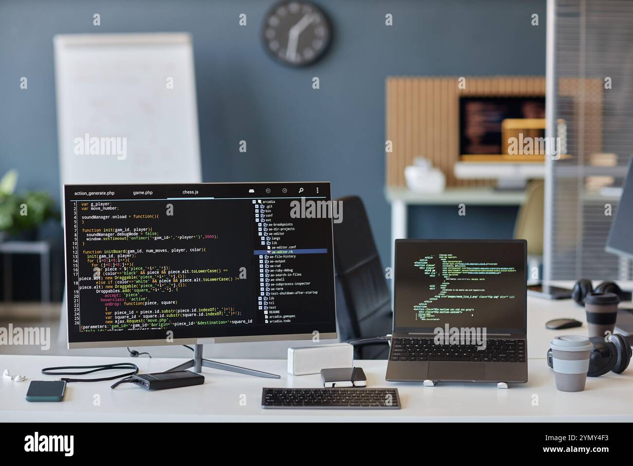 A modern office setup displaying desktop and laptop screens showing code. Background includes a clock and whiteboard, indicating a collaborative work Stock Photo