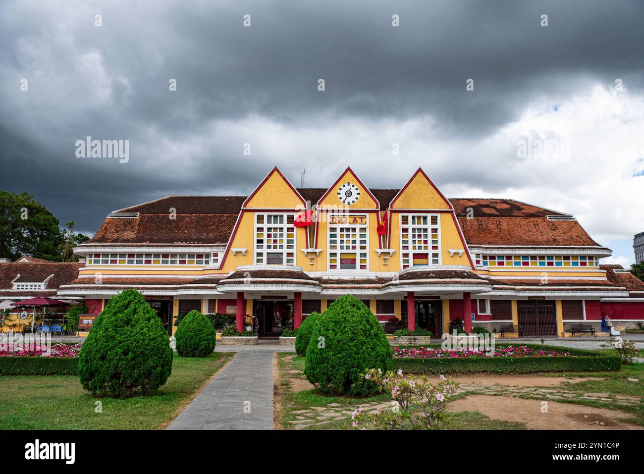 A yellow building with a clock on the front. The building has a red roof. There are several potted plants in front of the building Stock Photo