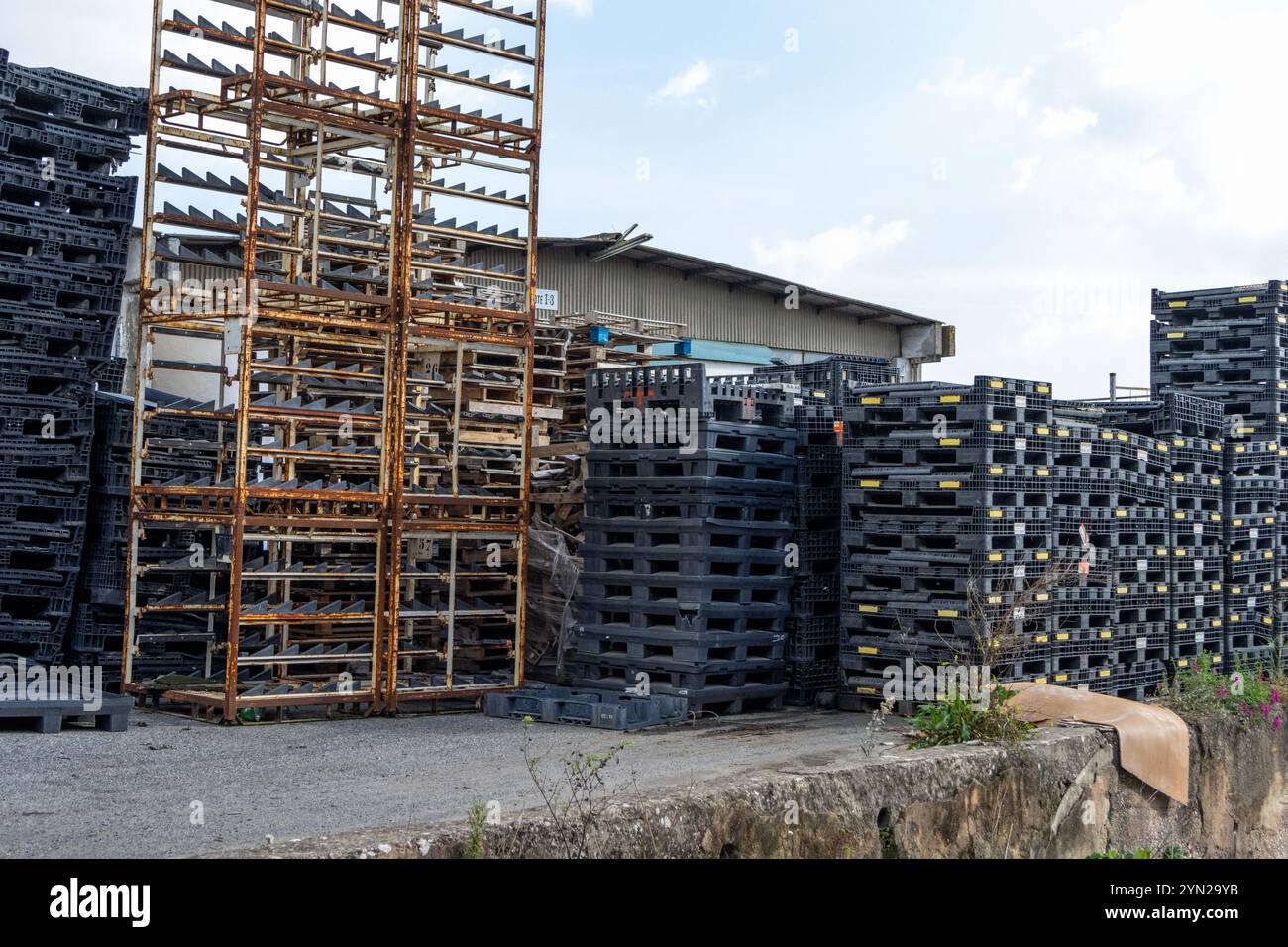 Stacks of black plastic pallets and rusty metal structures sit outside a warehouse, possibly indicating storage or logistics operations Stock Photo