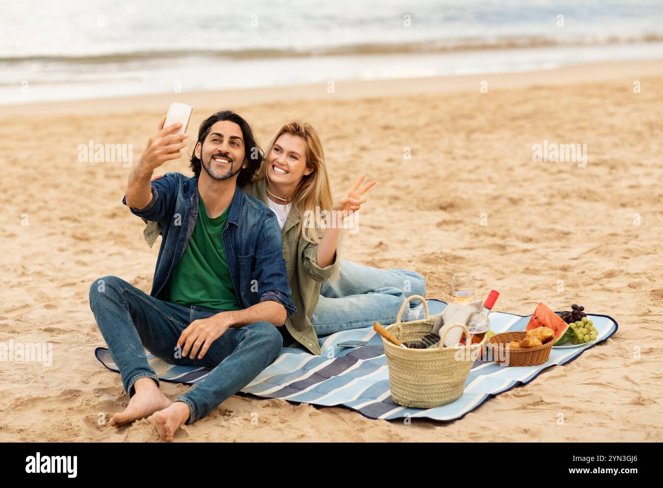 Couple enjoying a beach picnic while taking selfies during sunset Stock Photo
