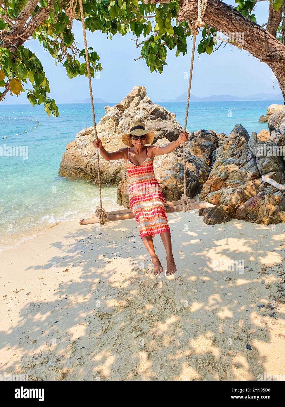 A person in a colorful dress swings gently from a hanging seat, surrounded by lush greenery and rocky shores, enjoying the warm sun and soft ocean breeze, Koh Kham Island Chonburi Samaesan Thailand Stock Photo