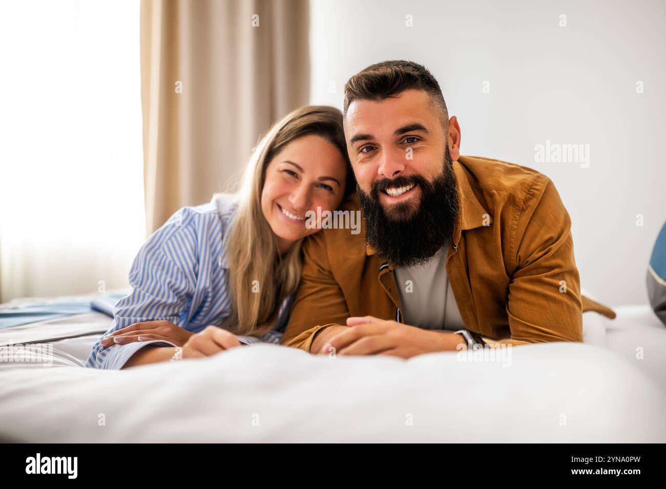 Young happy couple is lying on bed in their bedroom, looking at camera and smiling. Stock Photo