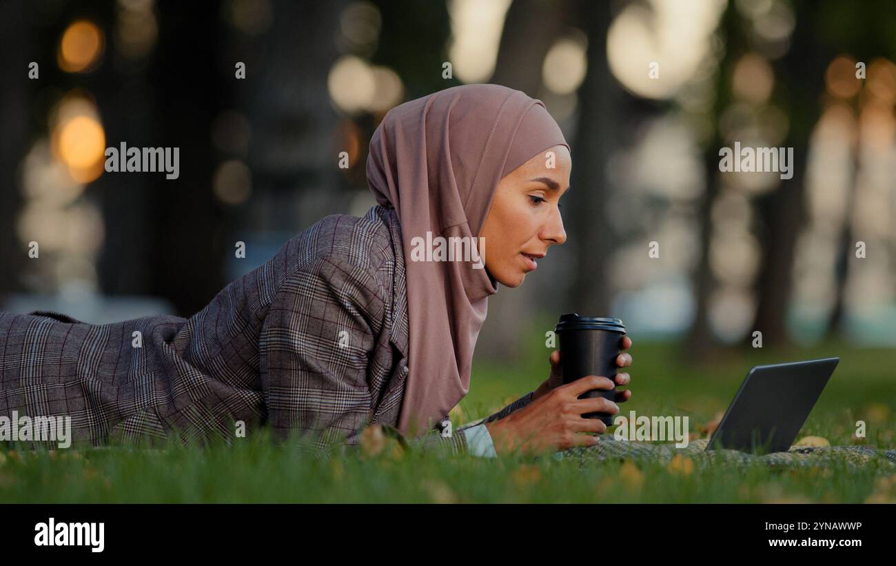 Muslim focused islamic girl business woman in hijab female student with laptop working online studying lying on grass green lawn in park drinking Stock Photo