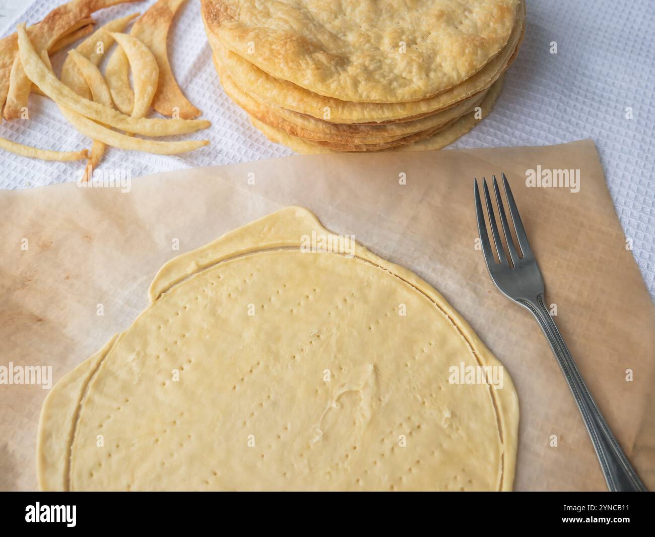 Raw layer rolled out on a baking paper and pierced with a fork, a pile of baked thin buttery layers, and dough scraps on a white waffle towel. Stock Photo