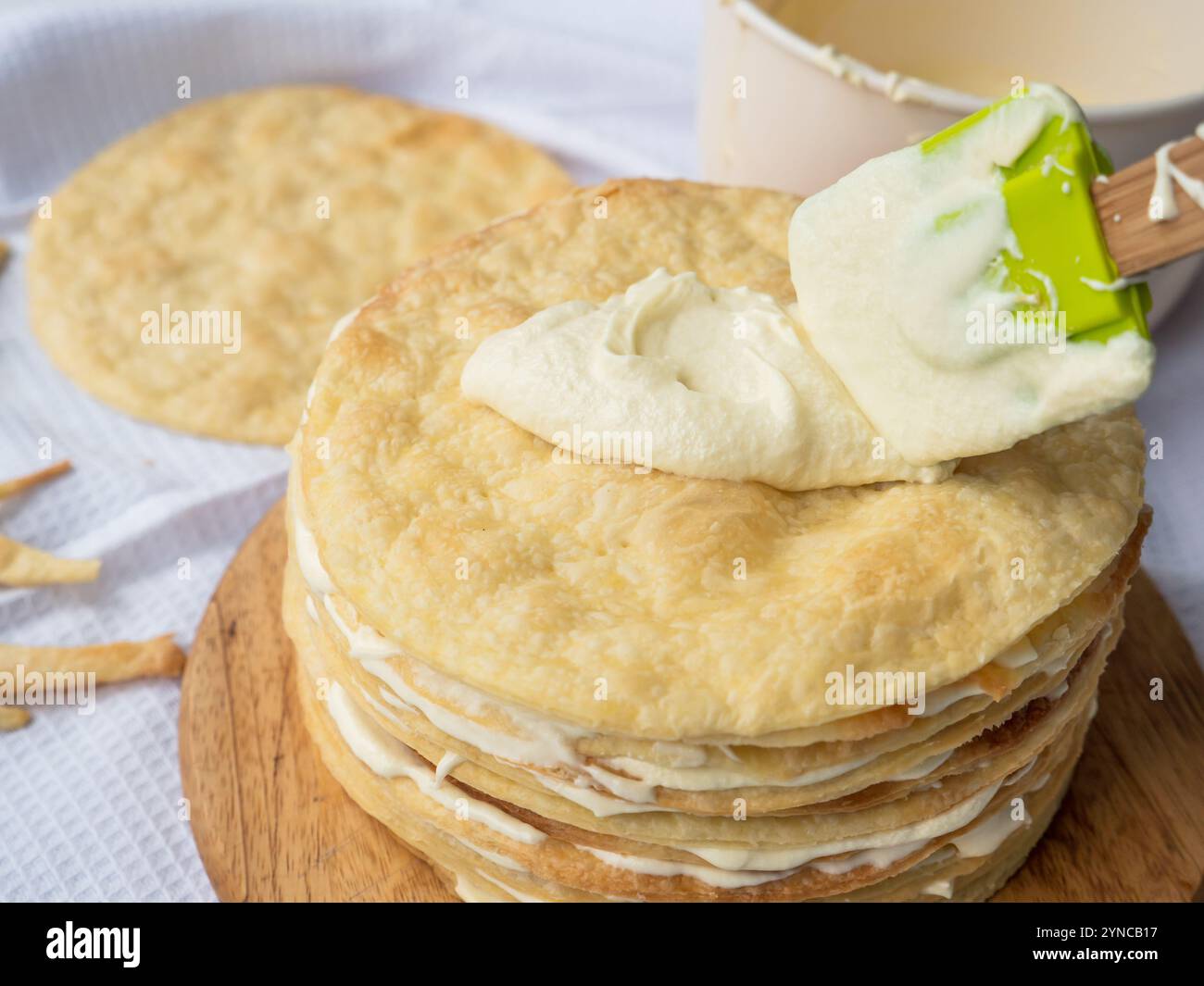 Silicone spatula spreading vanilla custard over a pile of baked thin round buttery layers on a wooden board, a bowl and a new pile waiting in back Stock Photo