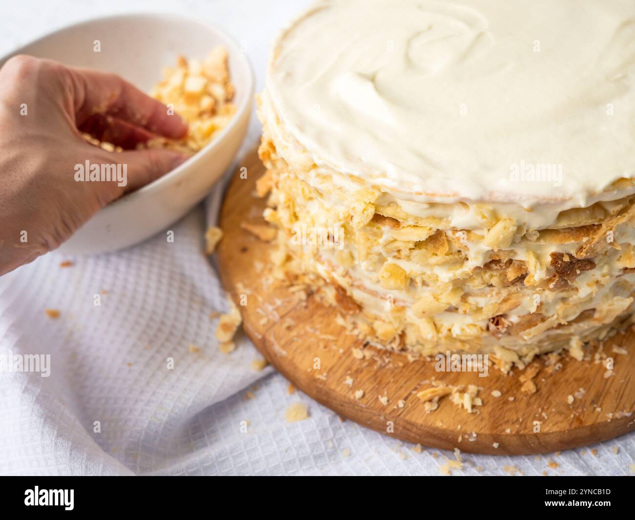 A hand sprinkling decorating crumbs over a pile of thin round layers spread with vanilla custard, a bowl of dough crumbs on a white waffle towel. Stock Photo
