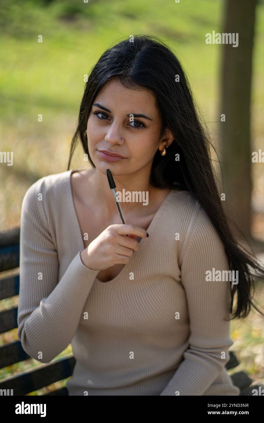 Young woman is sitting on a bench in a park, holding a pen and thinking Stock Photo