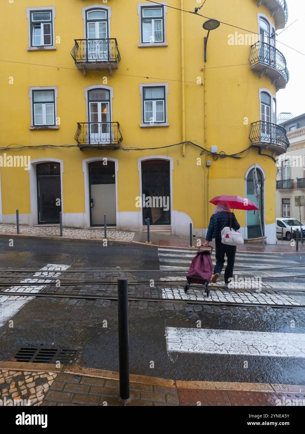 Older woman walking down the street with a red umbrella and shopping trolley on a rainy day. Yellow building in the background. Lisbon, Portugal - Smartphone Captured Stock Image