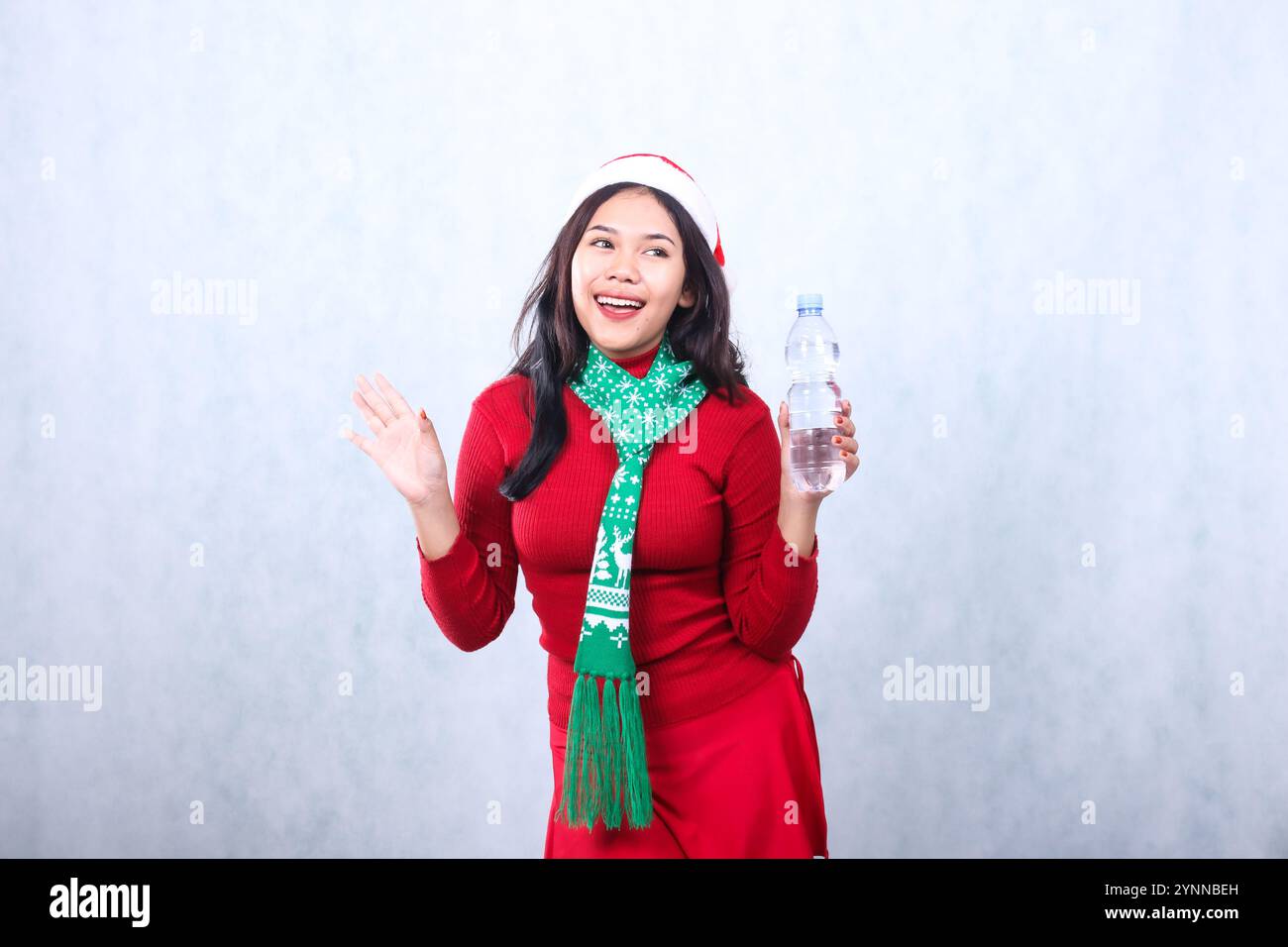 young beautiful Asian female wearing christmas sweater, Santa hat and scarf, cheerful left hand holding white water bottle and waving right, isolated Stock Photo